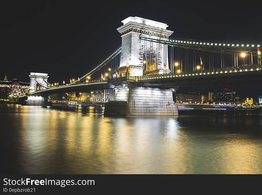 Stone stanchions of suspension bridge illuminated at night reflecting in river along urban waterfront. Stone stanchions of suspension bridge illuminated at night reflecting in river along urban waterfront.