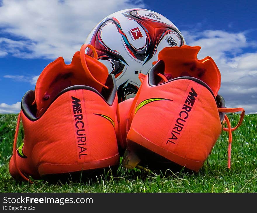 A close up of red soccer boots and a ball on green grass. A close up of red soccer boots and a ball on green grass.