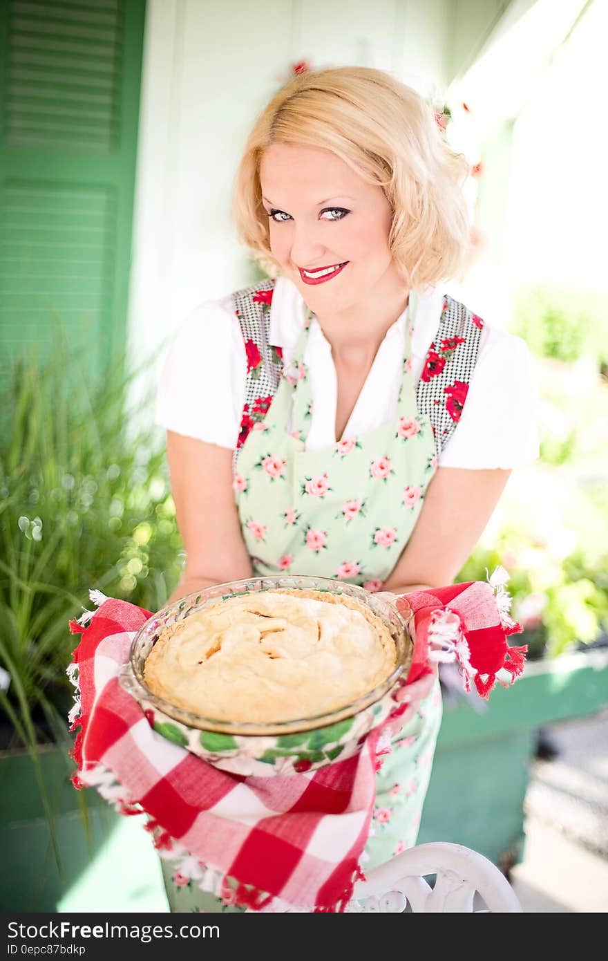 Woman in Pink White Floral Apron Smiling While Holding a White Creme Food during Daytime