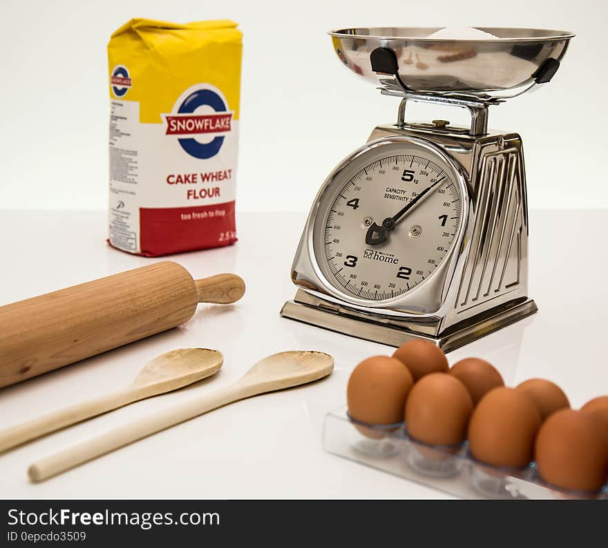 Weighing scales with flour in pan, tray of fresh brown eggs, packet of cake wheat flour, wooden spoons and rolling pin, white background. Weighing scales with flour in pan, tray of fresh brown eggs, packet of cake wheat flour, wooden spoons and rolling pin, white background.