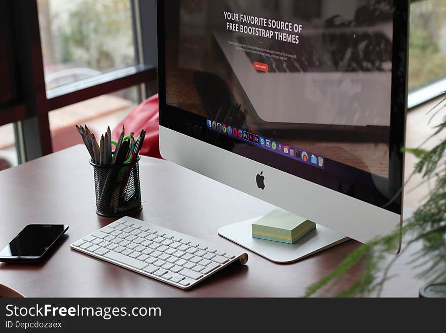 Large screen display and keyboard of Apple computer with memo pad, beaker filled with pencils and pens, office background. Large screen display and keyboard of Apple computer with memo pad, beaker filled with pencils and pens, office background.