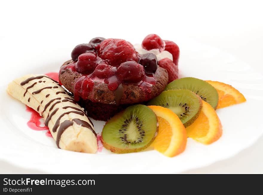 Banana Drizzled With Chocolate Syrup Beside Chocolate Cupcake With Raspberry and Kiwi and Orange Slices on the Side on White Plate