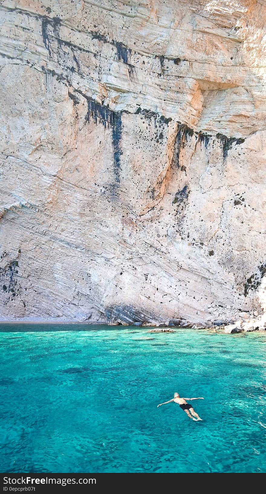 Man floating in blue waters along sheer cliff wall on sunny day. Man floating in blue waters along sheer cliff wall on sunny day.