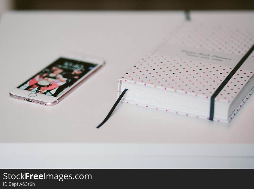 Rose Gold Iphone 6s Beside White and Black Polka Dots Book Both on Top of White Wooden Table