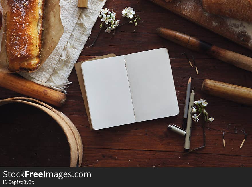 Still life of open blank book on table with pen and pencil, loaf of bread, burnt matches and flowers.