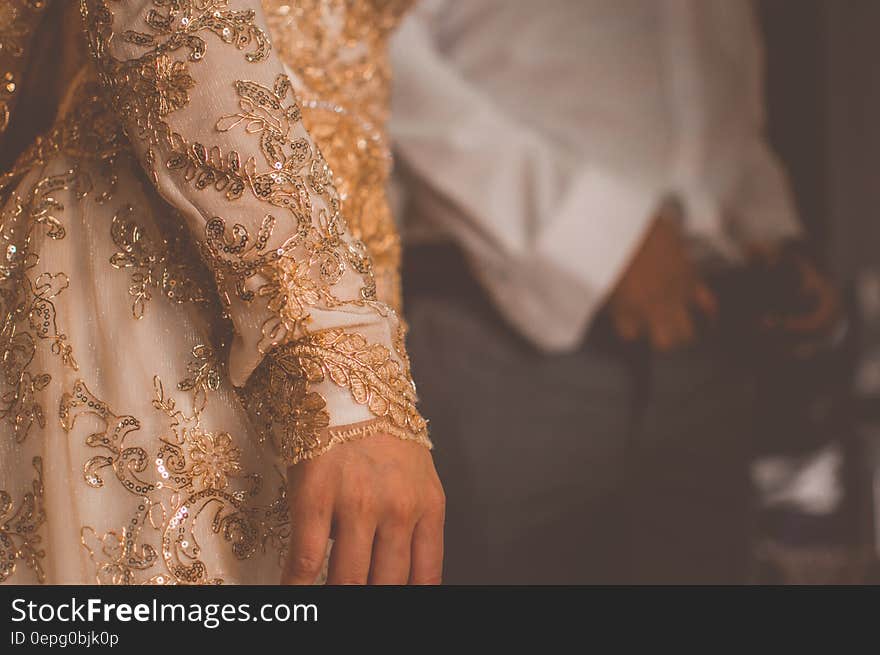 Close up of a woman wearing an elegant formal embroidered gown in gold and beige. Man in white shirt in the background. Close up of a woman wearing an elegant formal embroidered gown in gold and beige. Man in white shirt in the background.