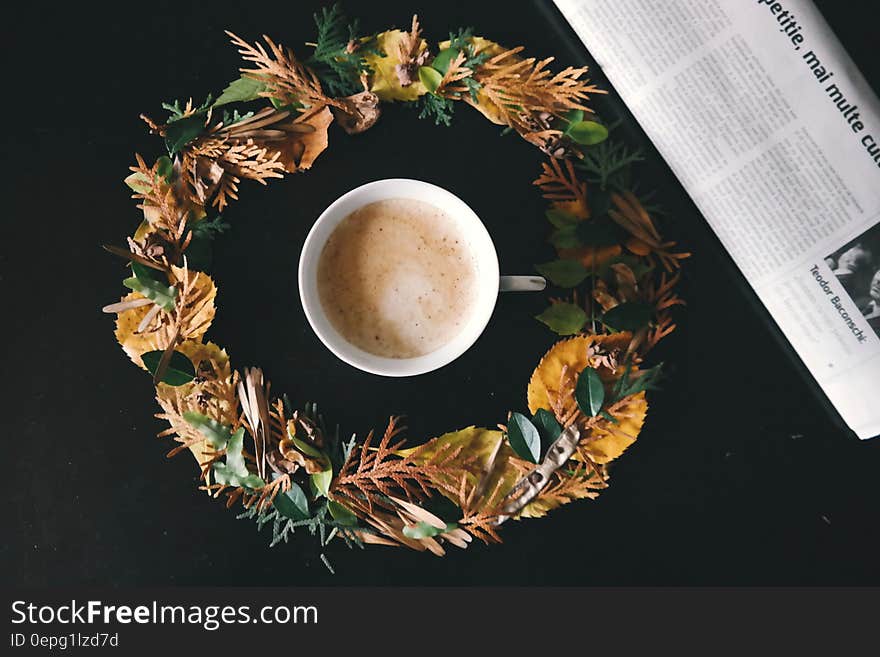 A cup of coffee in a wreath of leaves next to a newspaper. A cup of coffee in a wreath of leaves next to a newspaper.