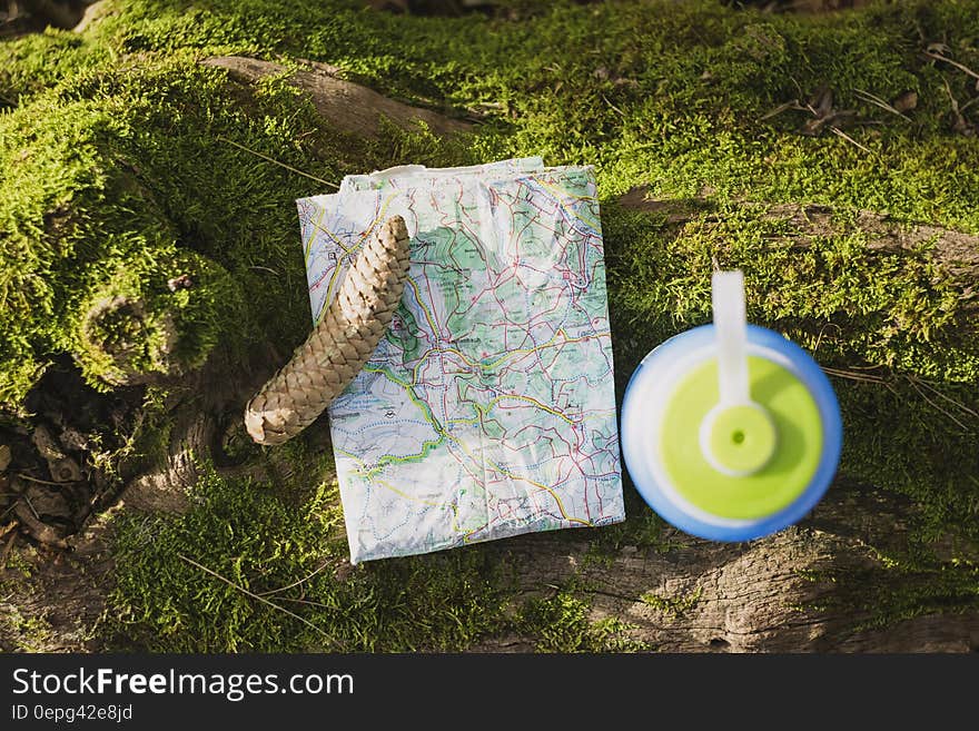 Map and pine cone with plastic water bottle on green lichen on sunny day.