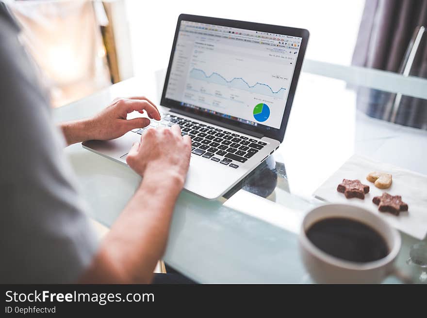 Man using laptop computer to analyze financial stock information on desk with coffee cup and cookies.