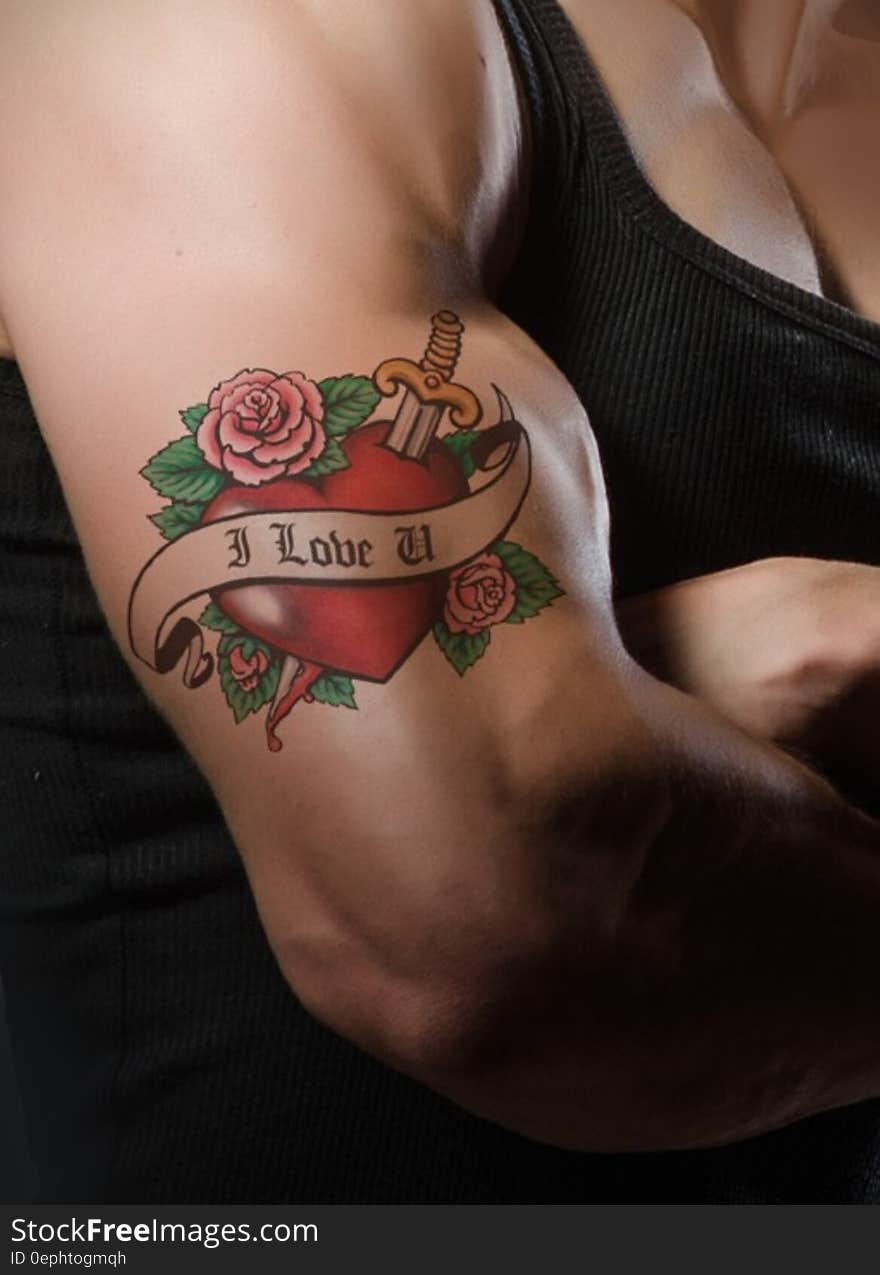 Tattoo on man's muscular arm with a picture of a red heart shape with roses and a dagger embellished with scroll and text "I love you", black background. Tattoo on man's muscular arm with a picture of a red heart shape with roses and a dagger embellished with scroll and text "I love you", black background.