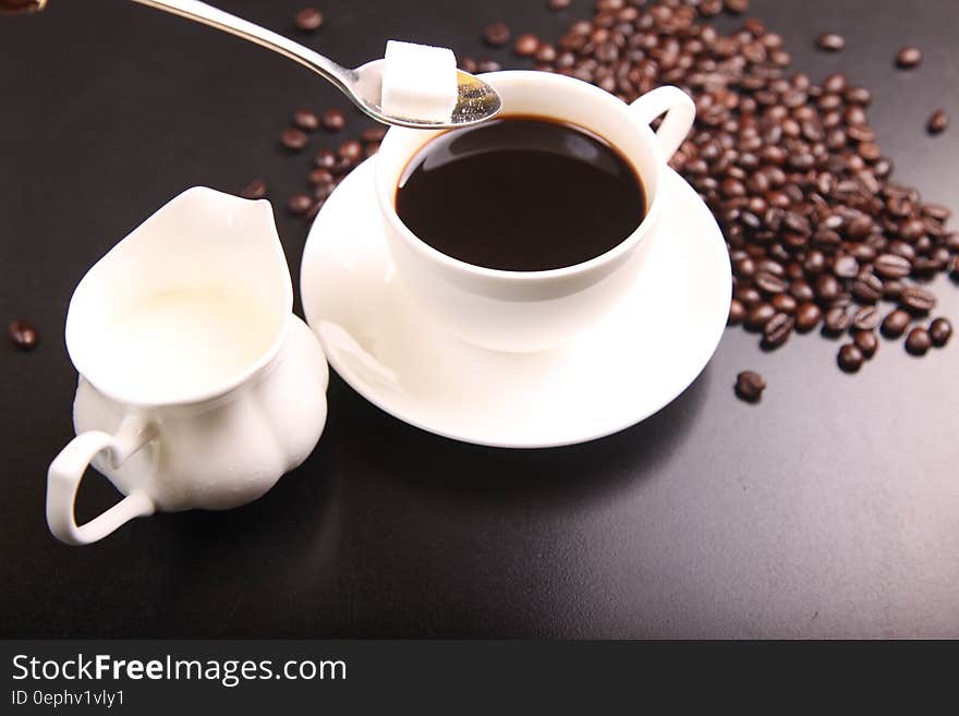 Cup of black coffee with spoon and sugar cube, cream jug and coffee beans laid out on plain dark table. Cup of black coffee with spoon and sugar cube, cream jug and coffee beans laid out on plain dark table.