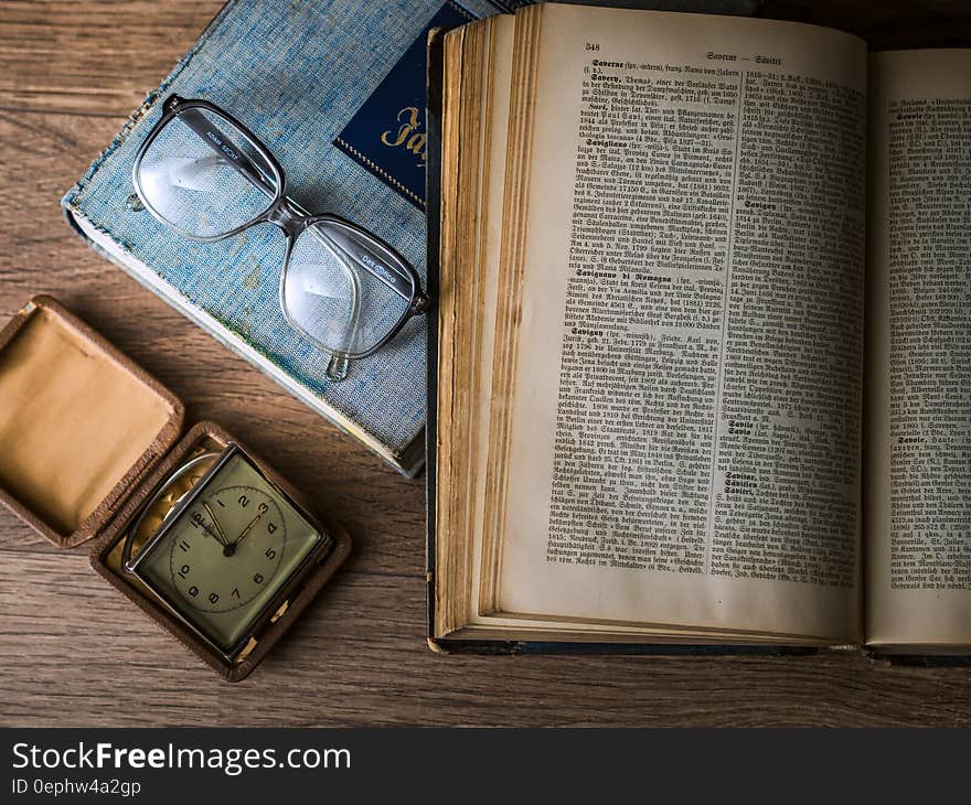 Eyeglasses next to an open old book and a small square clock beside. Eyeglasses next to an open old book and a small square clock beside.