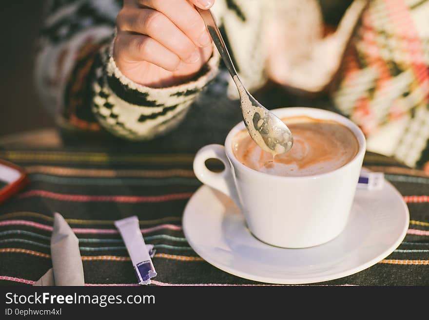 A close up of a cup of cappuccino, a hand stirring it with a teaspoon.