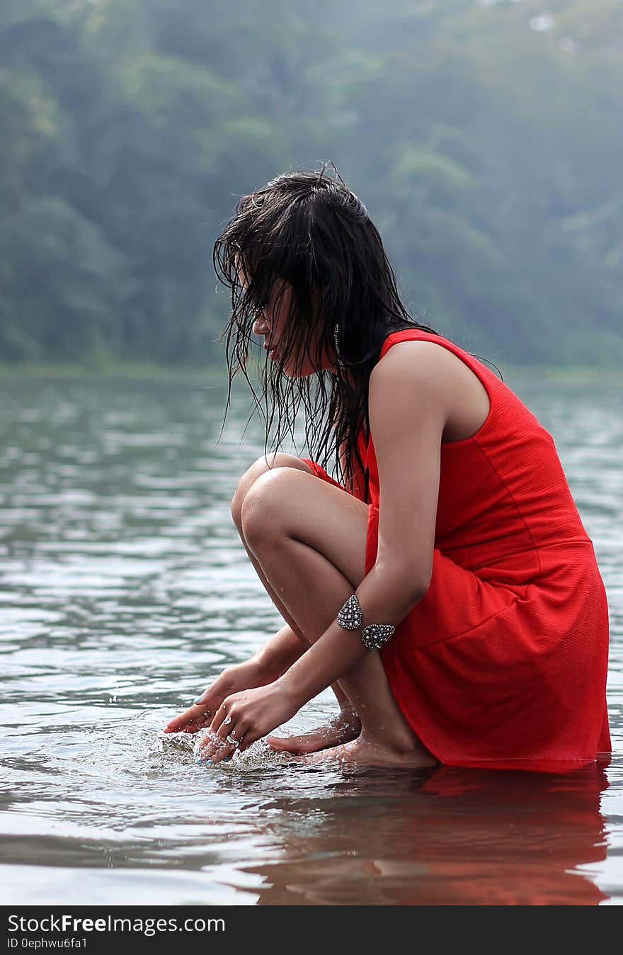 Side view of beautiful young woman in red dress with red hair crouching barefoot in lake. Side view of beautiful young woman in red dress with red hair crouching barefoot in lake.