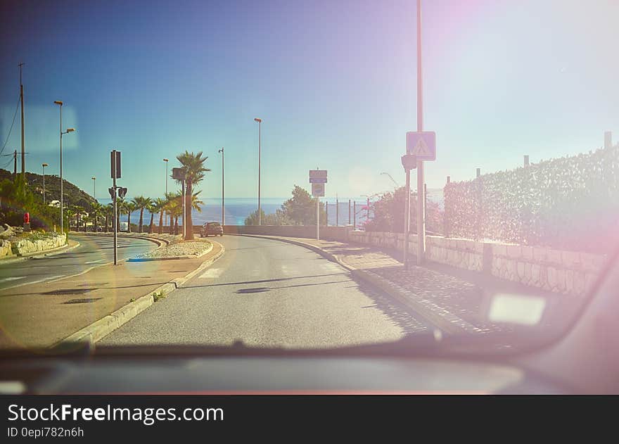 This picture is taken from inside a car through the windhield with the dashboard visible on the lower edge of the picture. The car is driving down a palm-lined ocean road under a blue sky with the ocean inthe background. The sunshine is reflected in the windshield of the car. This picture is taken from inside a car through the windhield with the dashboard visible on the lower edge of the picture. The car is driving down a palm-lined ocean road under a blue sky with the ocean inthe background. The sunshine is reflected in the windshield of the car.