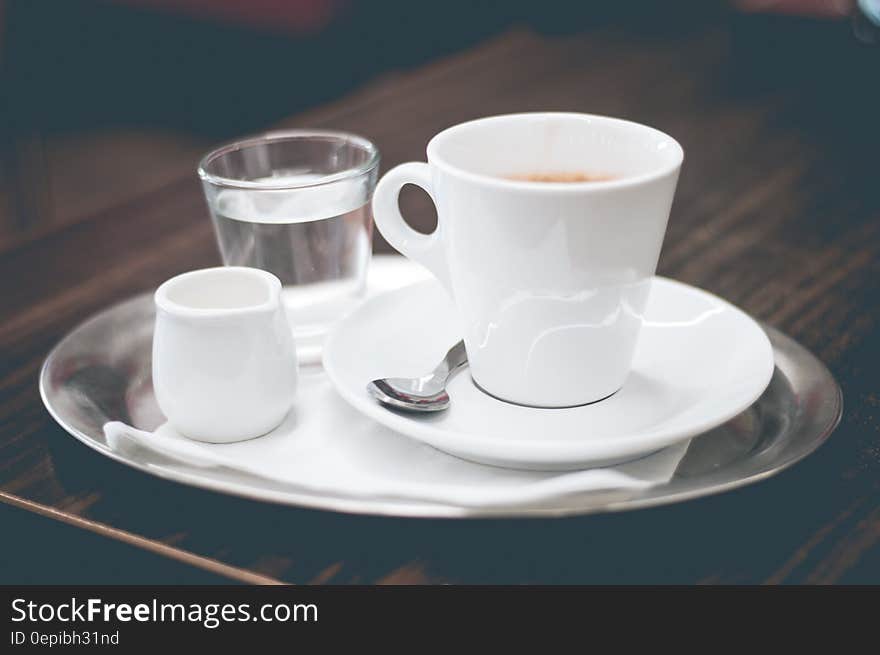 Coffee in a white China cup, cream in a jug, and a glass of water all on a silver tray, placed upon dark tray with wood grain. Coffee in a white China cup, cream in a jug, and a glass of water all on a silver tray, placed upon dark tray with wood grain.