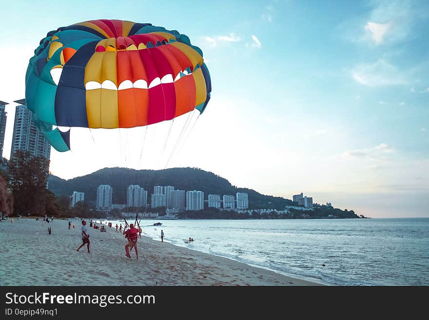 Skydiver and parachutist lands upon a sandy beach beside the sea with large colorful parachute still open, high rise apartments some distance away. Skydiver and parachutist lands upon a sandy beach beside the sea with large colorful parachute still open, high rise apartments some distance away.