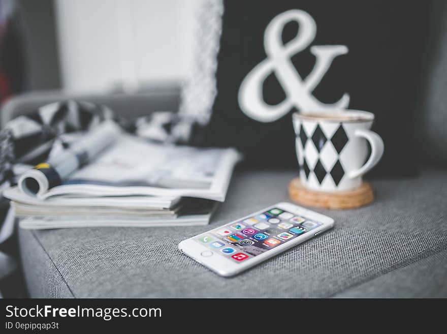 White mobile phone lying on a couch with cup and stack of magazines