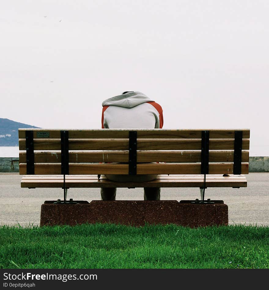 Person Wearing Grey and Orange Hoodie Sitting on Brown Wooden Park Bench during Daytime