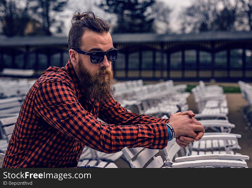 Man Wearing Black and Red Checkered Long Sleeve Shirt Wearing Black Wayfarer Sunglasses Sitting on White Wooden Chair