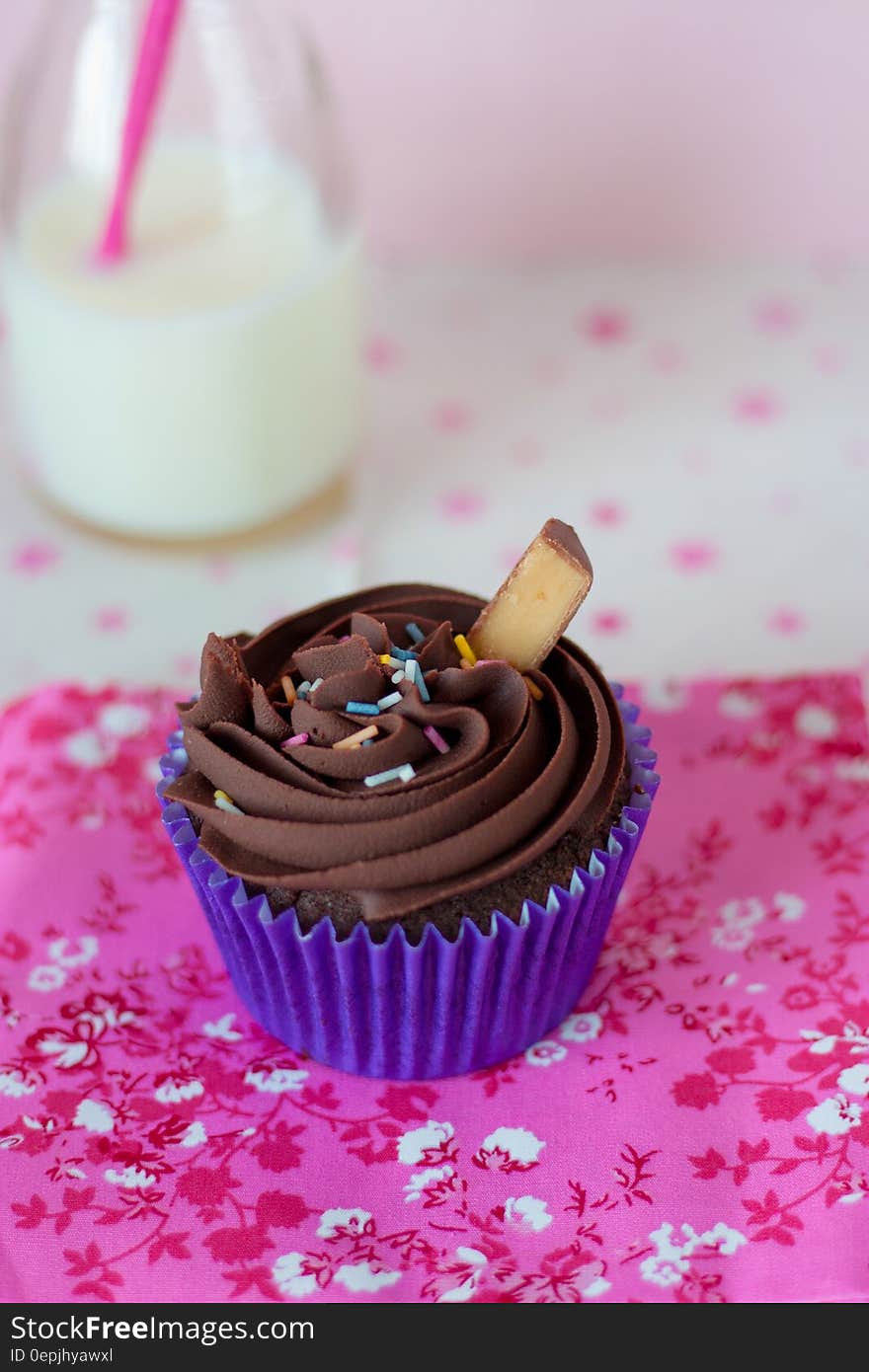 Close up of chocolate cupcake in blue wrapper with sprinkles on pink cloth with bottle of milk.