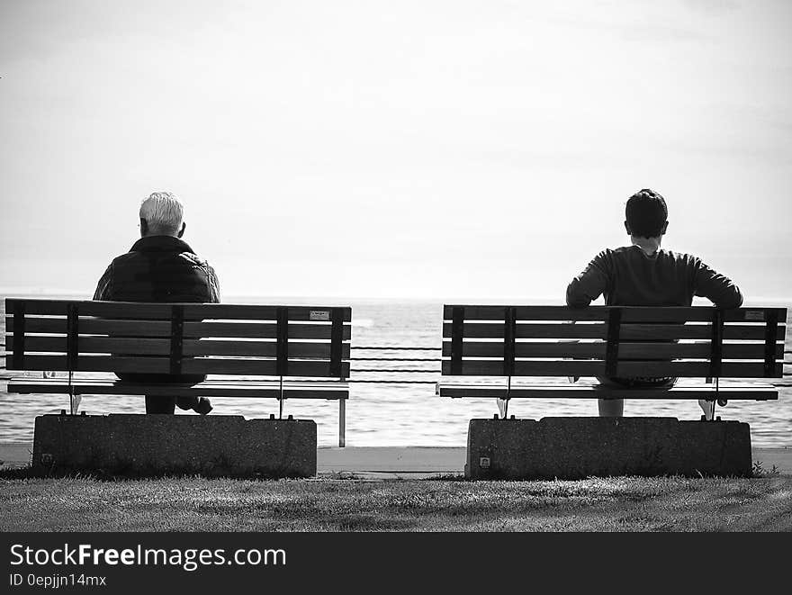 Grayscale Photo 2 Person Sitting in a Separate Benches on the Seaside