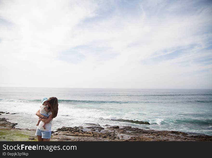 Mother Holding Her Baby in Front of Seashore during Daytime