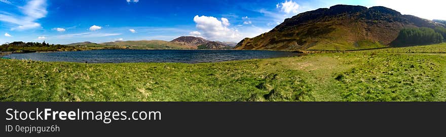 Panoramic view of countryside lake next to hills with blue sky and cloudscape.