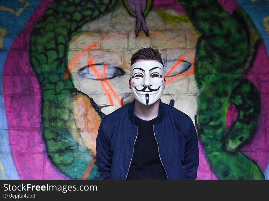 Man in White Mask in Black Crew Neck Shirt and Blue Zip Up Jacket Infront Graffiti Wall