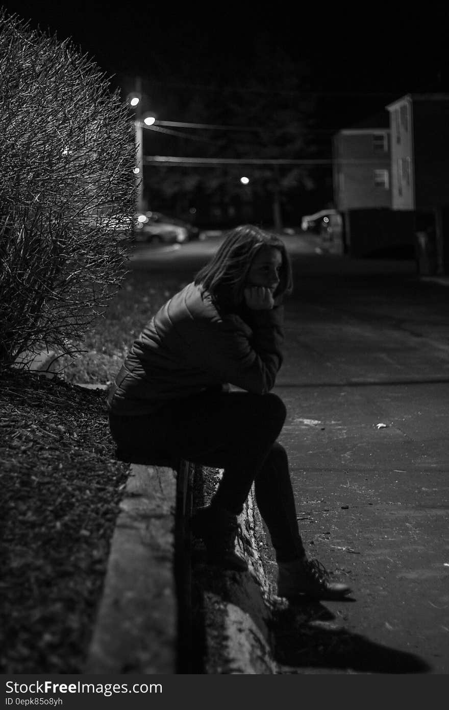 Woman Wearing Jacket Sitting on Concrete during Night Time