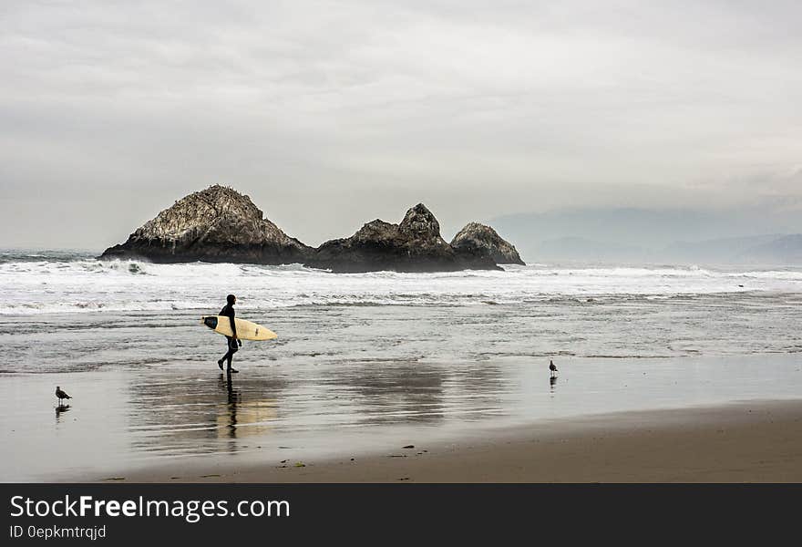 Man Carrying Surfboard in Front Bodies of Water on Brown Sand