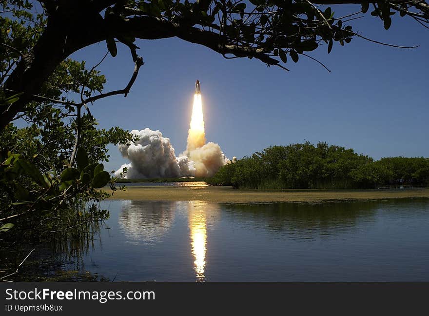 Rocket launch reflecting in calm waters of lake against blue skies.