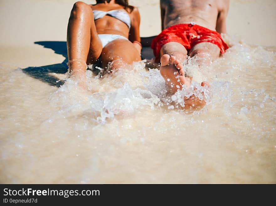 Anonymous couple on the beach both splashing the water with their feet. He is wearing red shorts and she a white bikini looking very sun tanned.