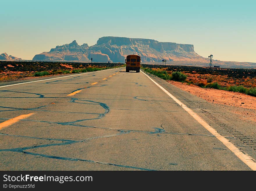 Road through red sandy desert with scrub growing along both sides and yellow bus approaching (or disappearing) . In distance is a table mountain and behind it a pale blue sky. Road through red sandy desert with scrub growing along both sides and yellow bus approaching (or disappearing) . In distance is a table mountain and behind it a pale blue sky.