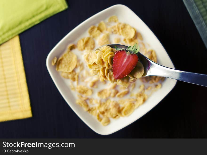 Spoon in white china bowl of cereal flakes in milk with sliced strawberry. Spoon in white china bowl of cereal flakes in milk with sliced strawberry.