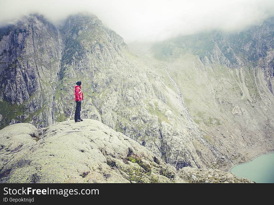 Character in red jacket and dark trousers standing on mountain top looking down upon blue Lake. Tops of nearby mountain range shrouded in fog and mist. Concept of getting to the top or great achievement. Character in red jacket and dark trousers standing on mountain top looking down upon blue Lake. Tops of nearby mountain range shrouded in fog and mist. Concept of getting to the top or great achievement.