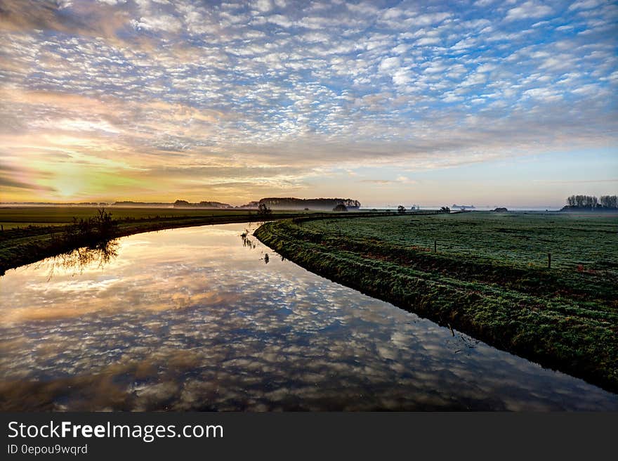 Clouds reflecting in river through green fields at sunset.