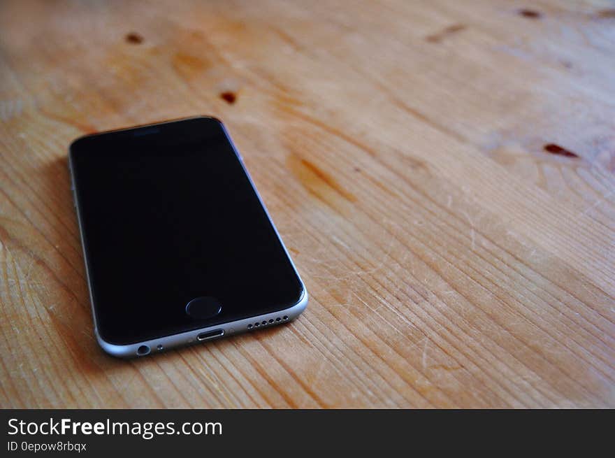 Apple iPhone with blank black screen and silver (or chrome) casing showing charger socket, speaker and headphone socket, lying upon grainy wooden table.