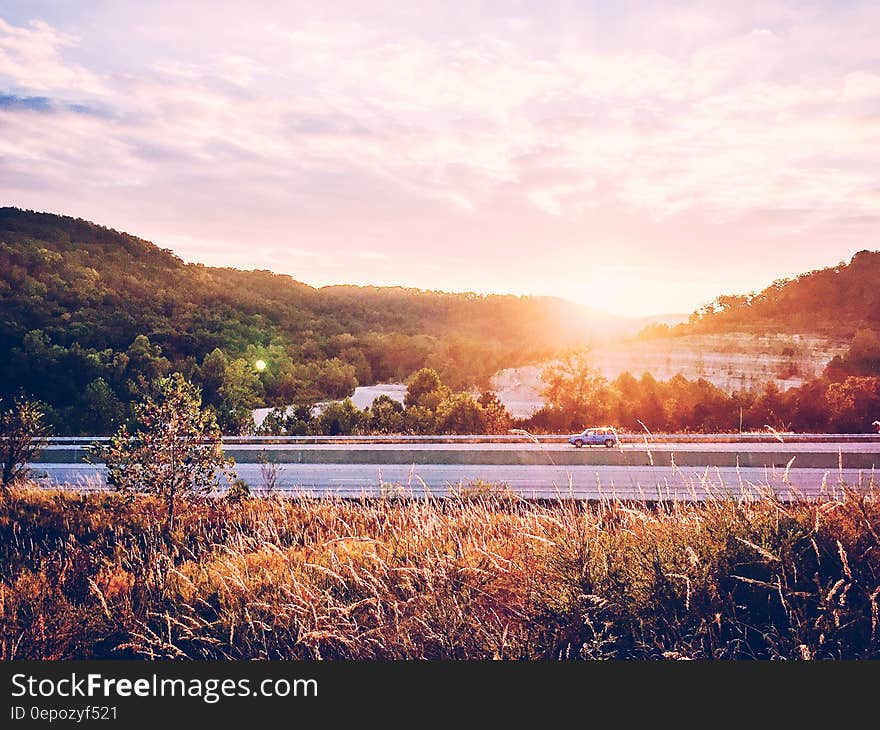 Sunrise over interstate highway with a single small car on the road and golden countryside with forests to left and right.