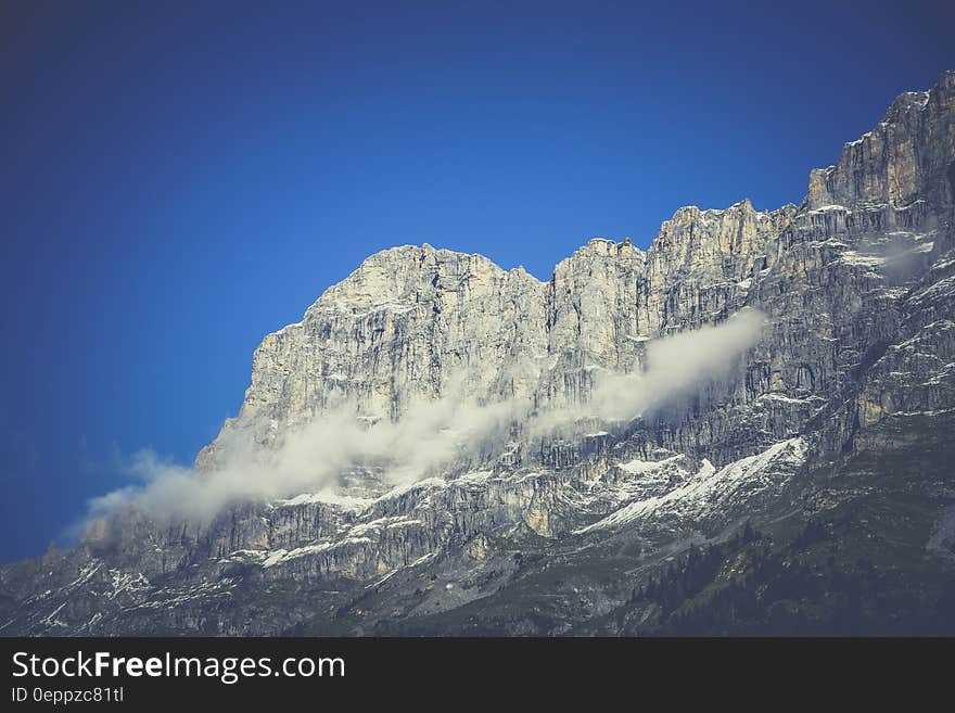 Steep jagged rocky cliff with sunlight illuminating detail and texture of the mountain, with mist and low cloud drifting by and blue sky above. Steep jagged rocky cliff with sunlight illuminating detail and texture of the mountain, with mist and low cloud drifting by and blue sky above.