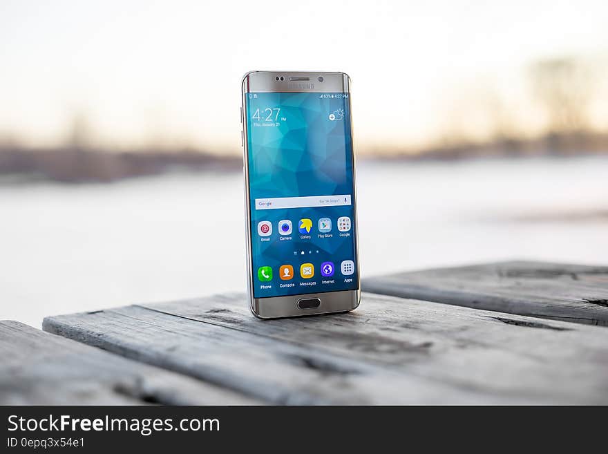 Samsung smartphone Galaxy S6 standing on the edge of a wooden jetty with screen illuminated and showing icons, background of sea or river beyond. Samsung smartphone Galaxy S6 standing on the edge of a wooden jetty with screen illuminated and showing icons, background of sea or river beyond.