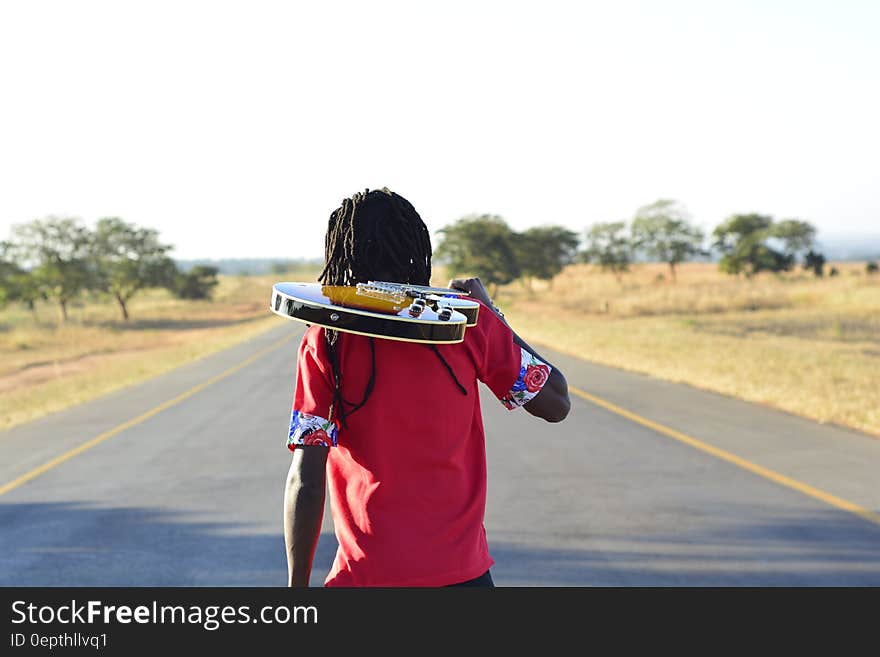 Person in Pink T Shirt Holding Brown Electric Guitar Walking on Blacktop Road during Daytime