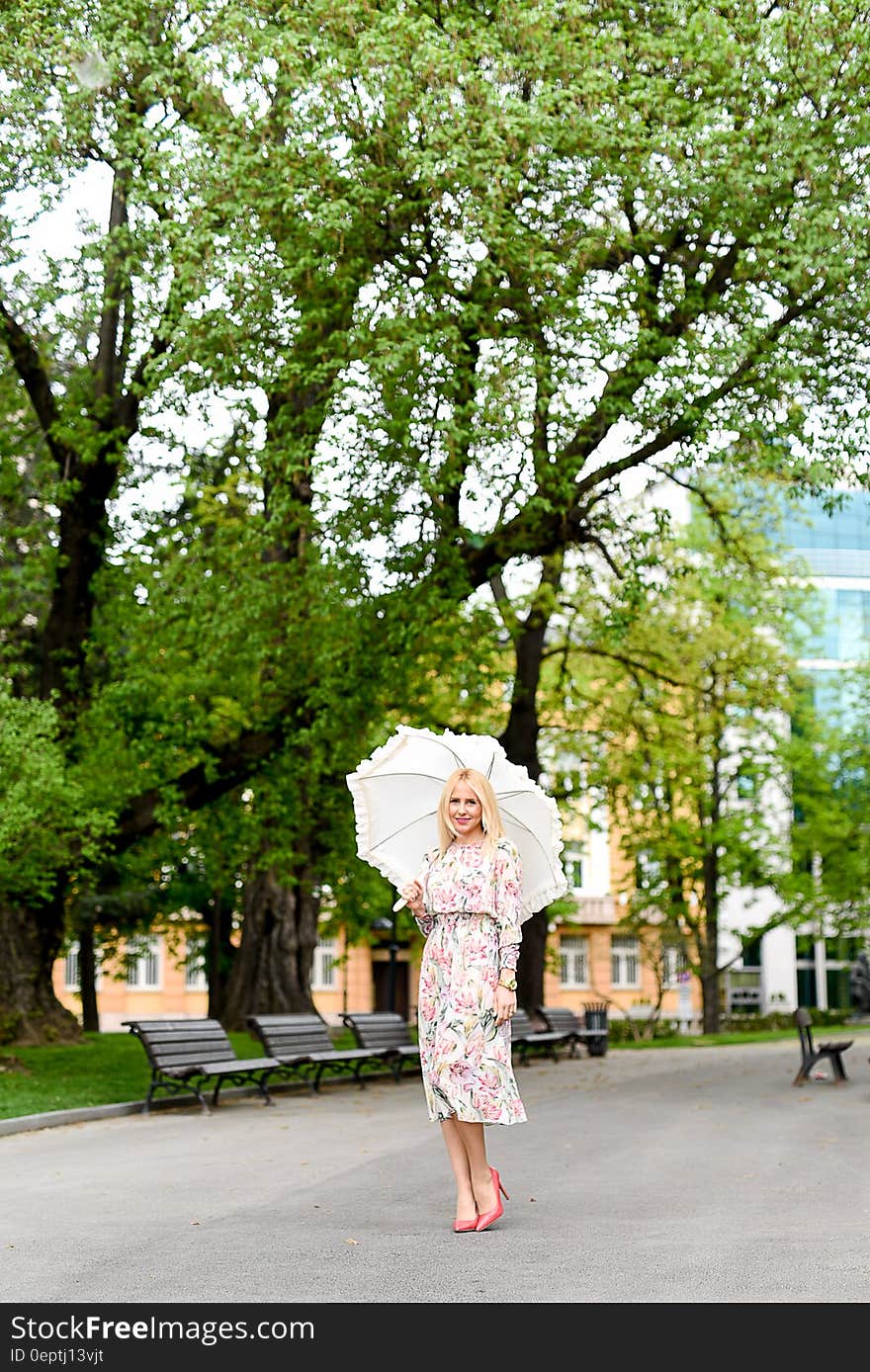 Woman in White Green and Red Floral Holding White Umbrella on Gray Concrete Pathway Near Brown Wooden Bench Near Trees during Dayt