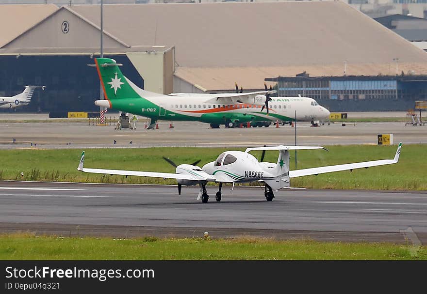 Airport with large green, white and red propeller driven passenger aircraft and smaller white private plane on the runway, hangars in the background. Airport with large green, white and red propeller driven passenger aircraft and smaller white private plane on the runway, hangars in the background.