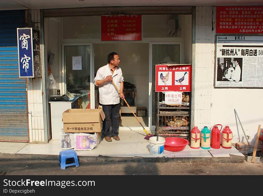 Asian shopkeeper sweeping storefront on sunny day.