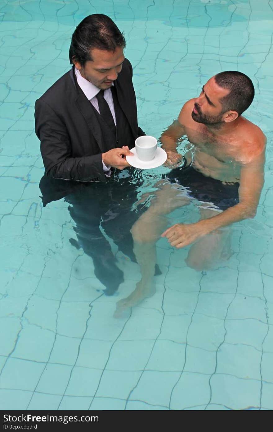 Man with business suit holding cup of coffee in swimming pool next to man in swimming trunks. Man with business suit holding cup of coffee in swimming pool next to man in swimming trunks.