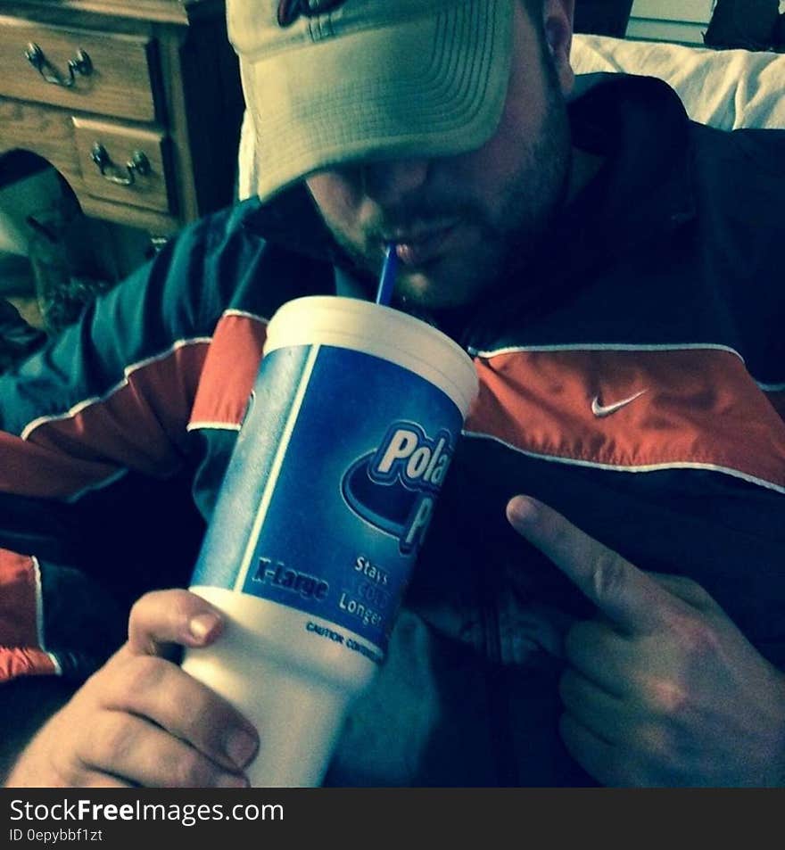 Young man with baseball cap shading face at home drinking fizzy drink through straw. Young man with baseball cap shading face at home drinking fizzy drink through straw.