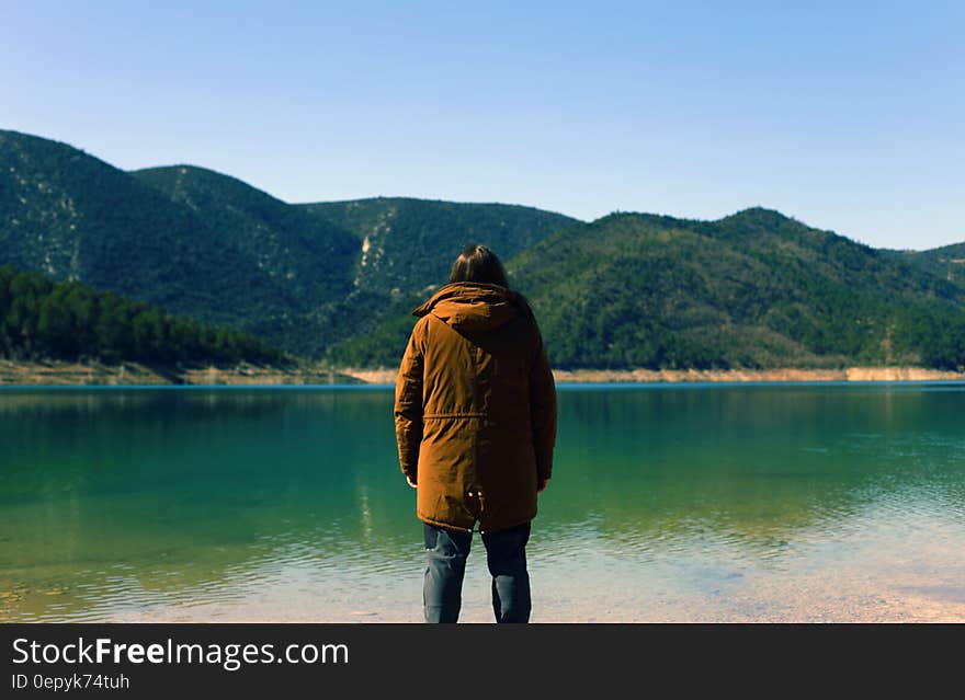 Man wearing brown jacket with hood, dark trousers and boots looking across tranquil lake towards distant mountains. Man wearing brown jacket with hood, dark trousers and boots looking across tranquil lake towards distant mountains.