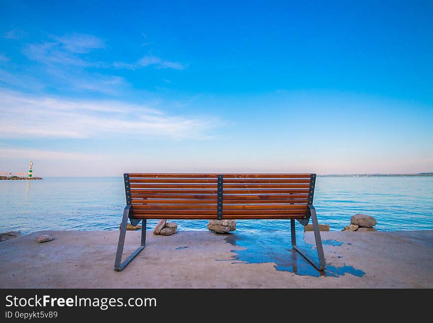 Empty bench on waterfront against blue skies on sunny day.