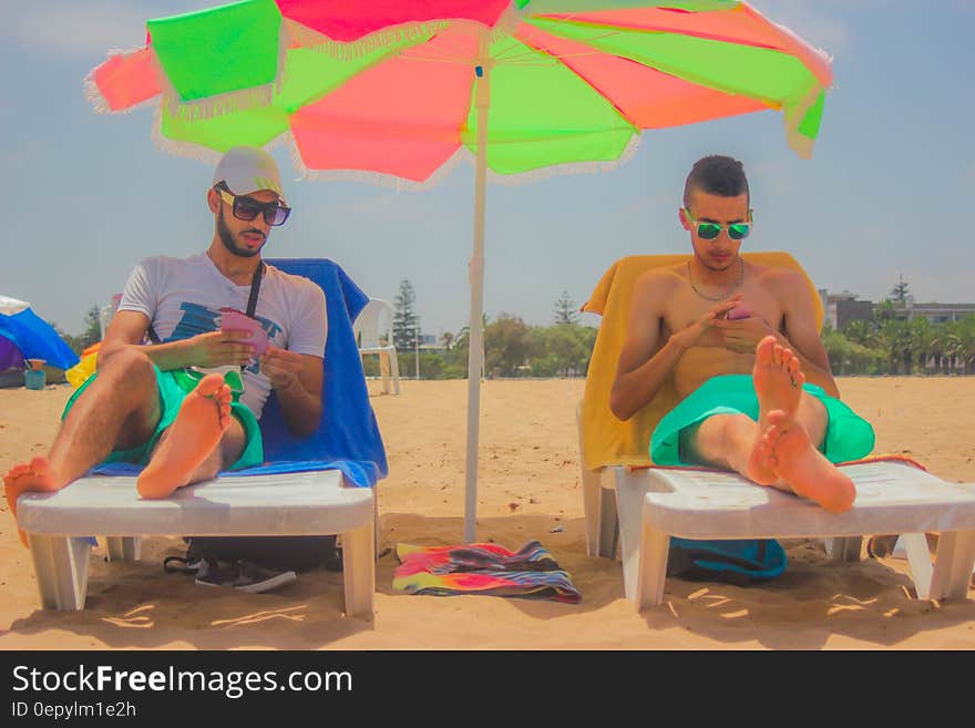 2 Man Sitting on White Beach Lounger With Green and Pink Patio Umbrella in Between